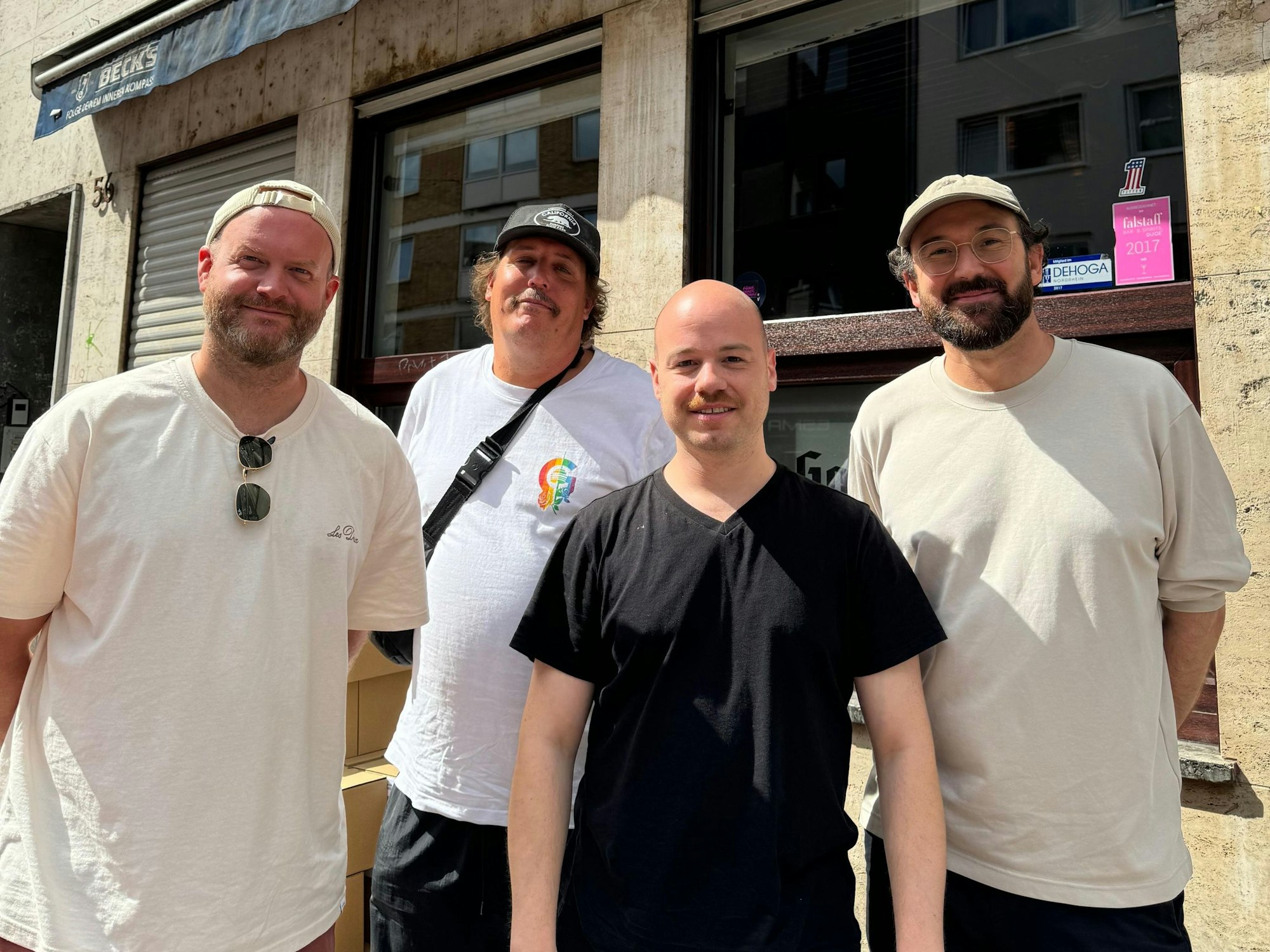 Vier Männer stehen vor einer Kneipe in Köln und lächeln in die Kamera.
