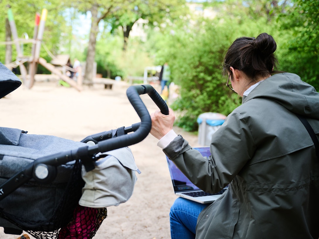 Eine Mutter sitzt am Laptop, während sie neben dem Kinderwagen auf dem Spielplatz sitzt.