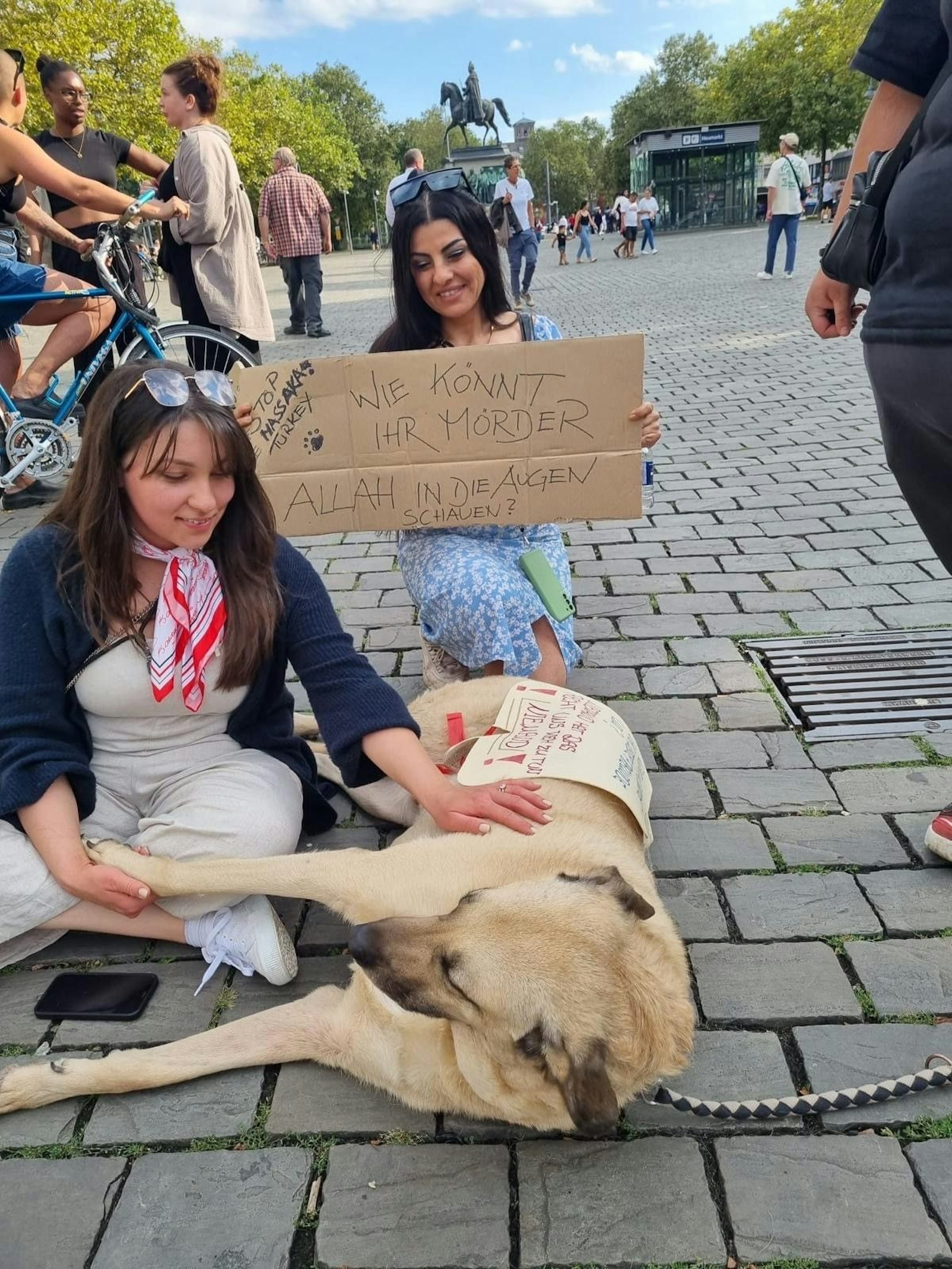 Zwei Frauen sitzen mit einem Hund auf einem Platz bei einer Demo.