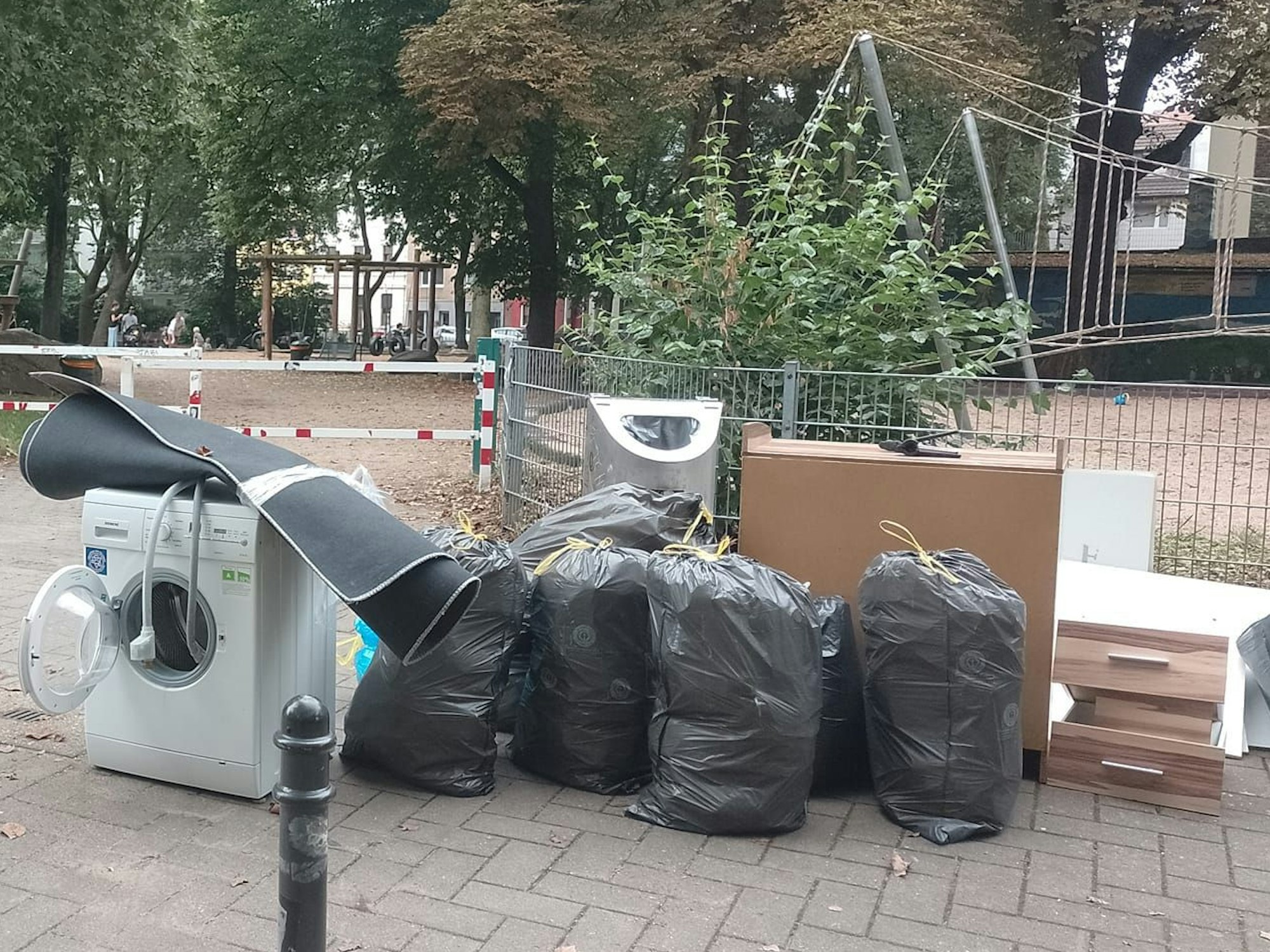 Am Zaun eines Spielplatzes stehen mehrere Müllsäcke, eine Waschmaschine sowie Sperrmüll.