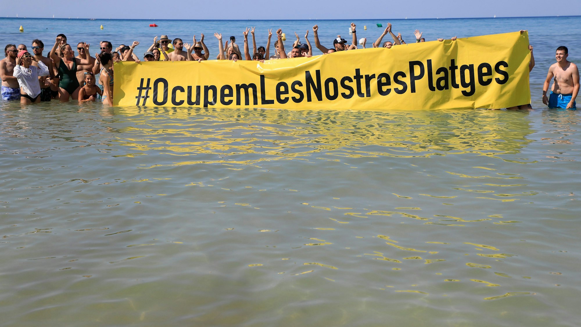 Leute halten an einem Strand auf Mallorca ein Banner „#Besetzen wir unsere Strände“ aus Protest gegen den Massentourismus auf der Balearen-Insel.