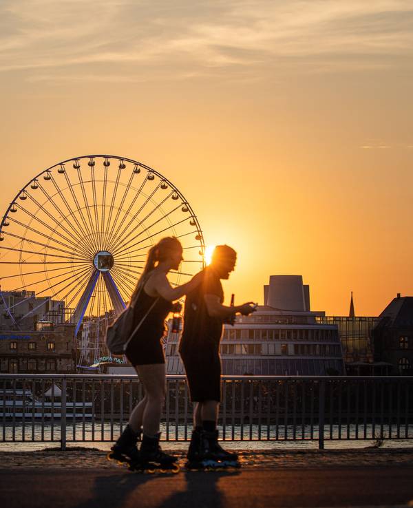 Die Sonne geht am Abend hinter dem Riesenrad am Rheinauhafen unter während im Vordergrund zwei Menschen auf Inline-Skates auf der Deutzer Werft unterwegs sind.
