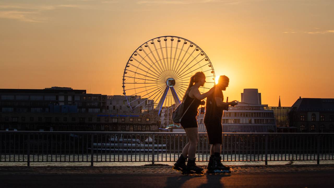 Die Sonne geht am Abend hinter dem Riesenrad am Rheinauhafen unter während im Vordergrund zwei Menschen auf Inline-Skates auf der Deutzer Werft unterwegs sind.