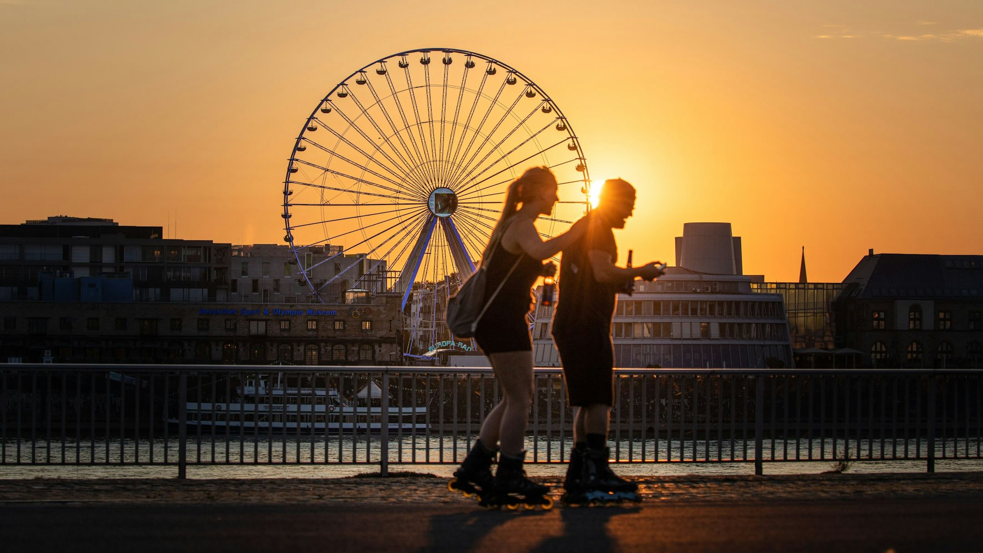 Die Sonne geht am Abend hinter dem Riesenrad am Rheinauhafen unter während im Vordergrund zwei Menschen auf Inline-Skates auf der Deutzer Werft unterwegs sind.