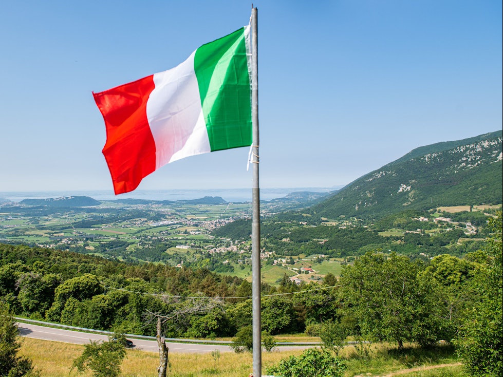Eine italienische Flagge weht auf einem Balkon, in der Ferne ist der Gardasee zu sehen. Kriminelle in Italien haben Hunderte Menschen mit der Gründung eines fiktiven Staats und damit vermeintlich verbundenen Vorteilen um insgesamt Hunderttausende von Euro betrogen. (zu dpa "Betrüger ergaunern mit erfundenem Antarktisstaat rund 400.000 Euro ") +++ dpa-Bildfunk +++