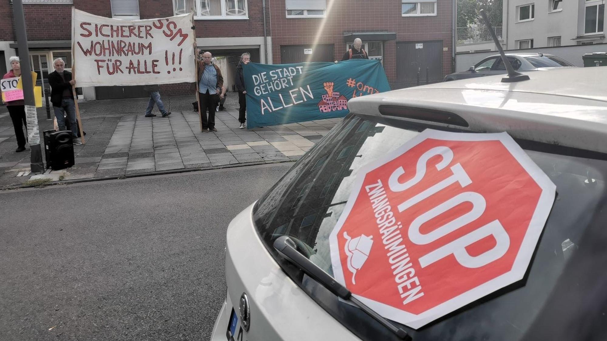 Vor einem Haus stehen mehrere Menschen mit Bannern, darauf steht unter anderem „Sicherer Wohnraum für alle!!!“ An der Rückscheibe eines Autos lehnt ein Schild mit der Aufschrift: „Stop Zwangsräumungen“.