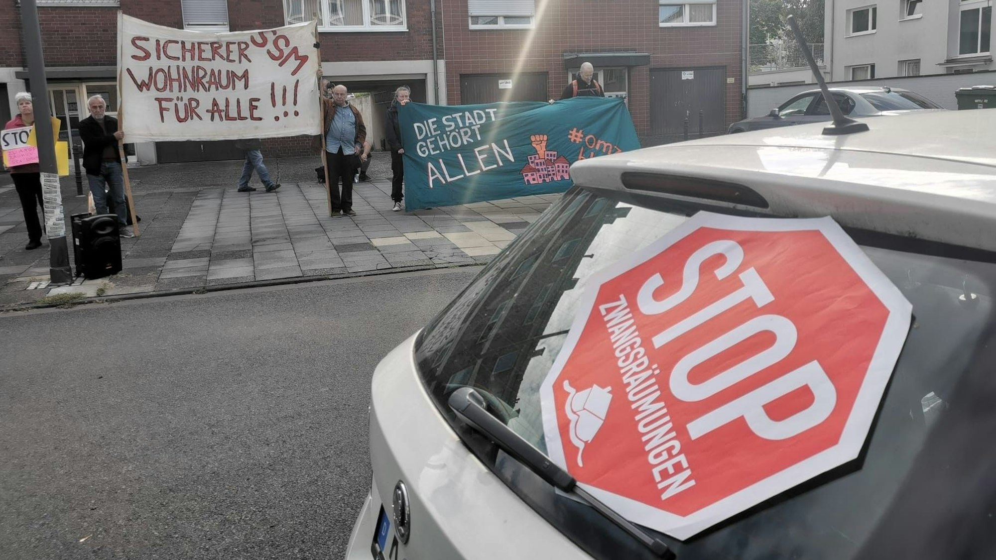 Vor einem Haus stehen mehrere Menschen mit Bannern, darauf steht unter anderem „Sicherer Wohnraum für alle!!!“ An der Rückscheibe eines Autos lehnt ein Schild mit der Aufschrift: „Stop Zwangsräumungen“.