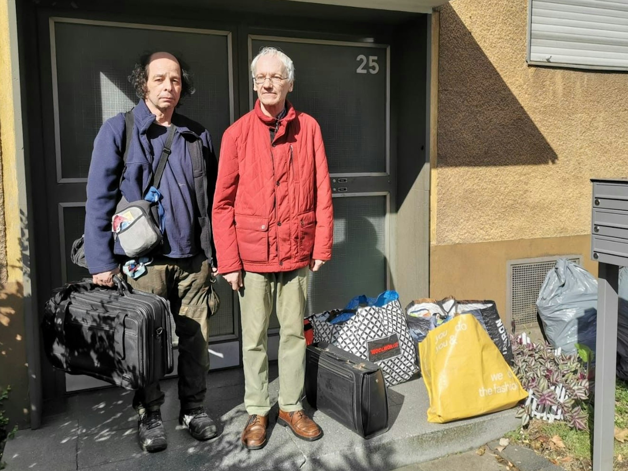 Zwei ältere Männer stehen mit Koffern vor einer Haustür.
