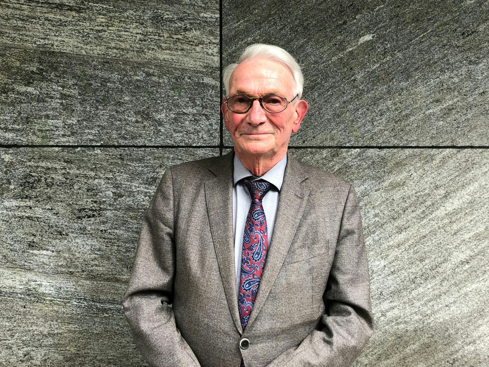 Hansmanfred Boden steht im Anzug vor einer Wand und schaut in die Kamera.