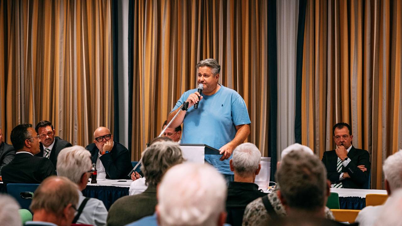 Ein Mann in blauem Shirt steht an einem Rednerpult und spricht in ein Mikrofon.