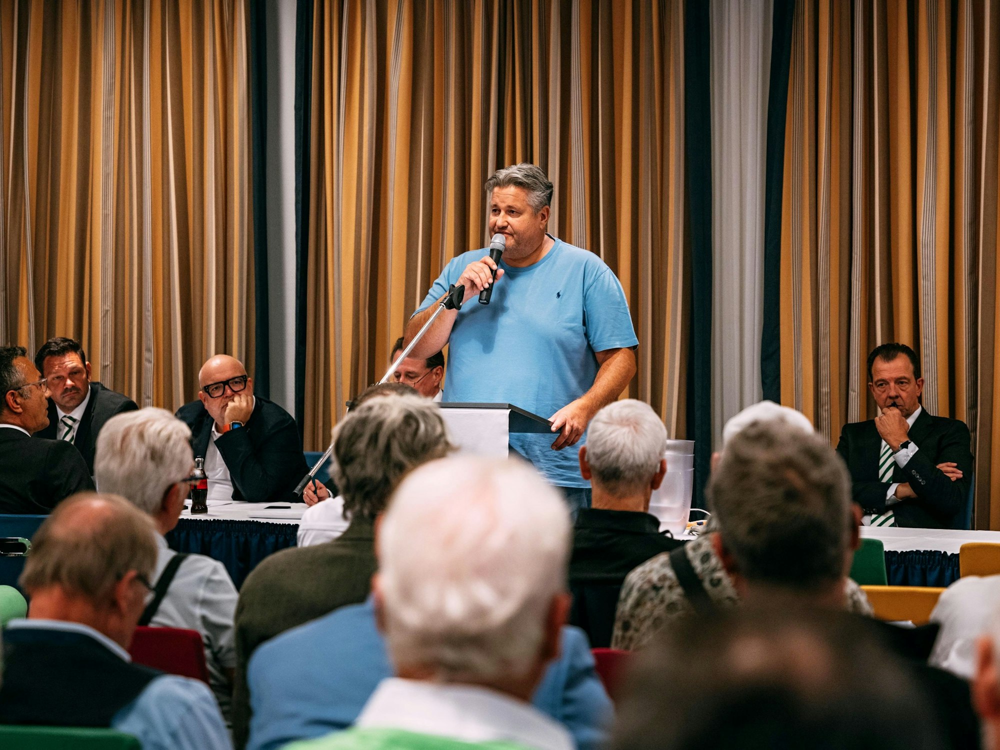 Ein Mann in blauem Shirt steht an einem Rednerpult und spricht in ein Mikrofon.