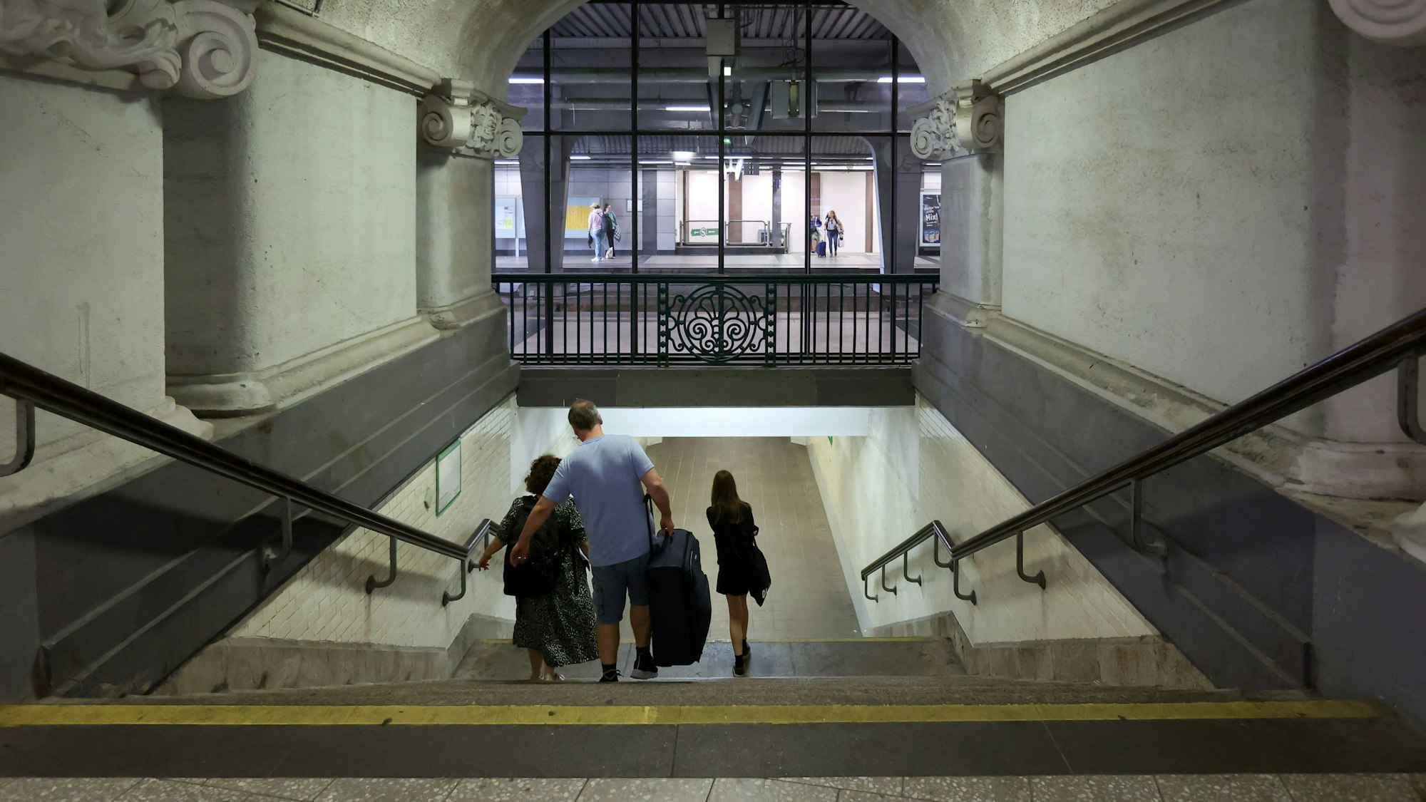 Drei Menschen gehen im Bahnhof Köln Messe/Deutz mit ihrem Gepäck eine Treppe herunter.