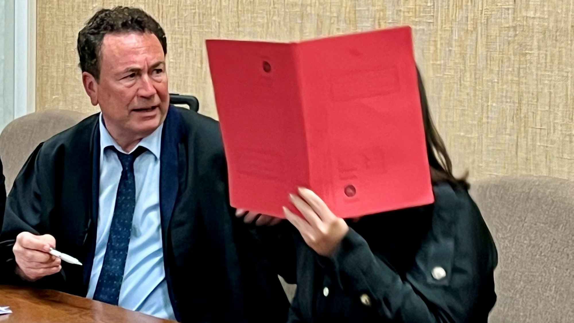 Eine Frau sitzt neben ihrem Anwalt und hält sich eine Mappe vor das Gesicht.