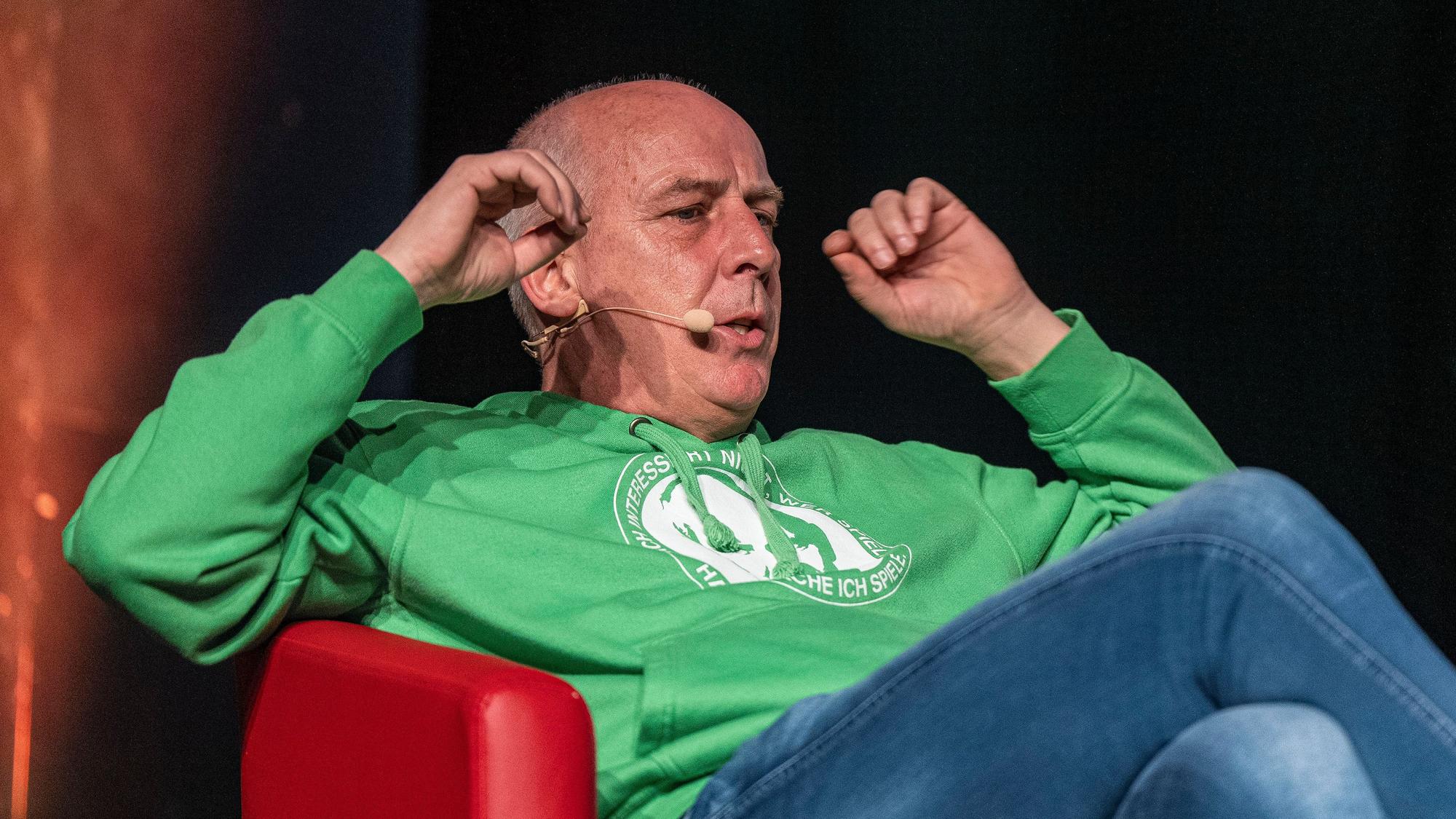 Mario Basler sitzt bei einem Auftritt des „Doppelpass on Tour“ im Sessel und gestikuliert.