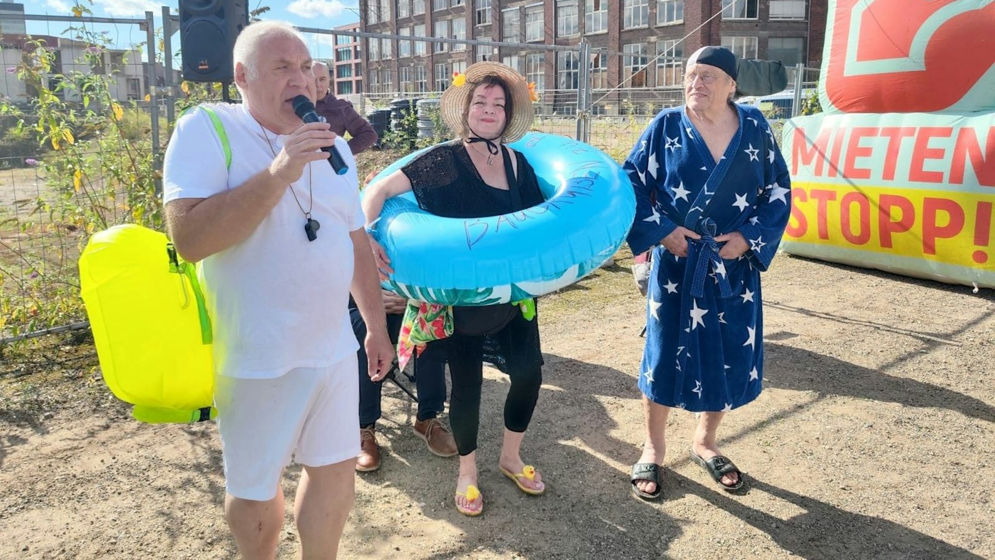 Ein Mann in kurzer Hose spricht ins Mikrofon, neben ihm eine Frau mit einem Schwimmring und ein Mann im Bademantel.