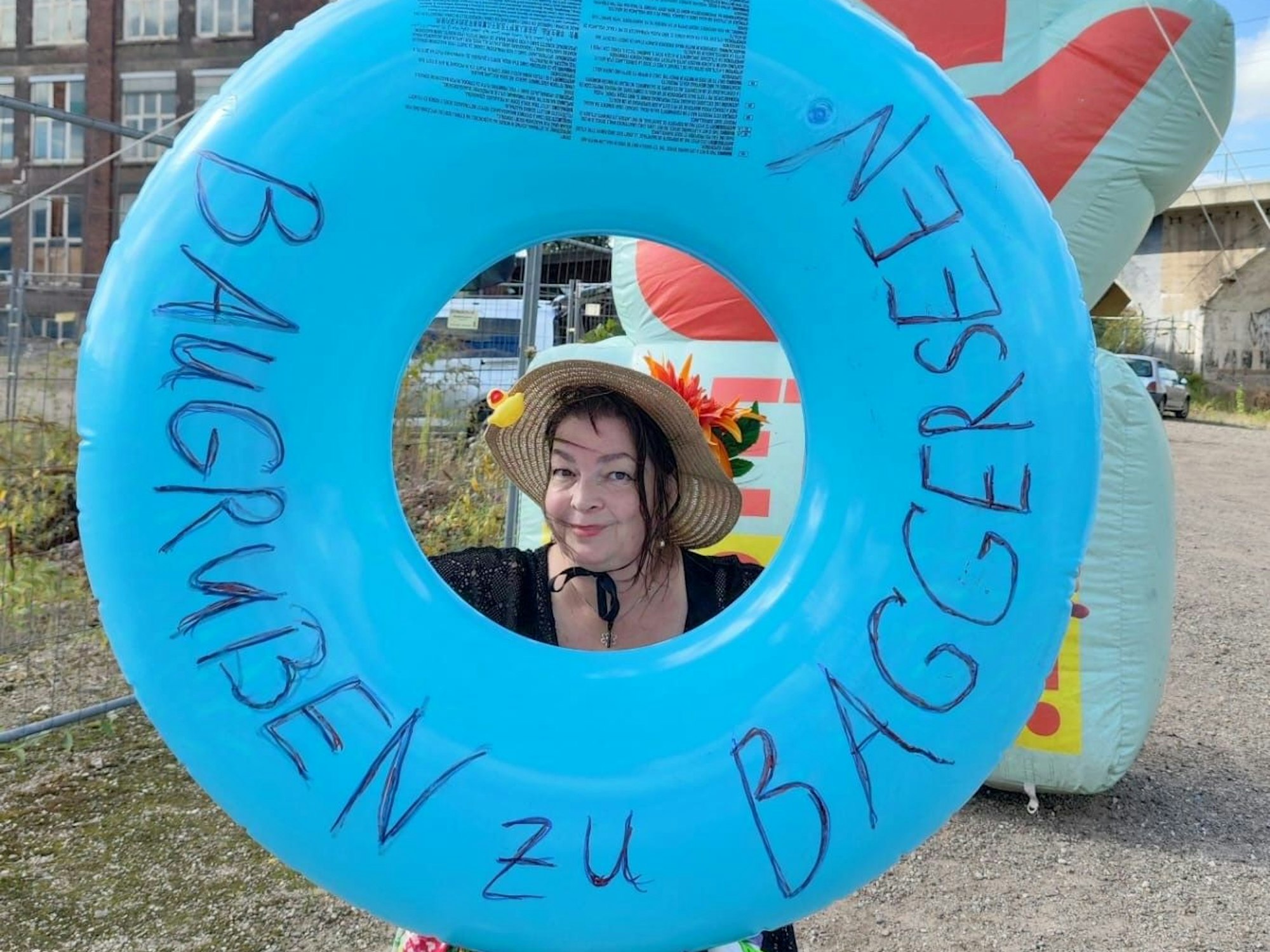 Eine Frau guckt durch das Loch eines großen Schwimmrings, auf dem „Baugruben zu Baggerseen“ geschrieben steht.