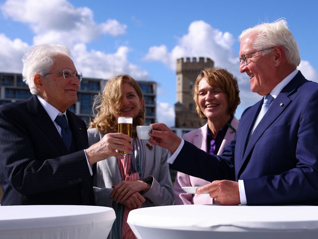 Zwei Männer stoßen mit einem Bier beziehungsweise einem Espresso an, dahinter stehen zwei Frauen.