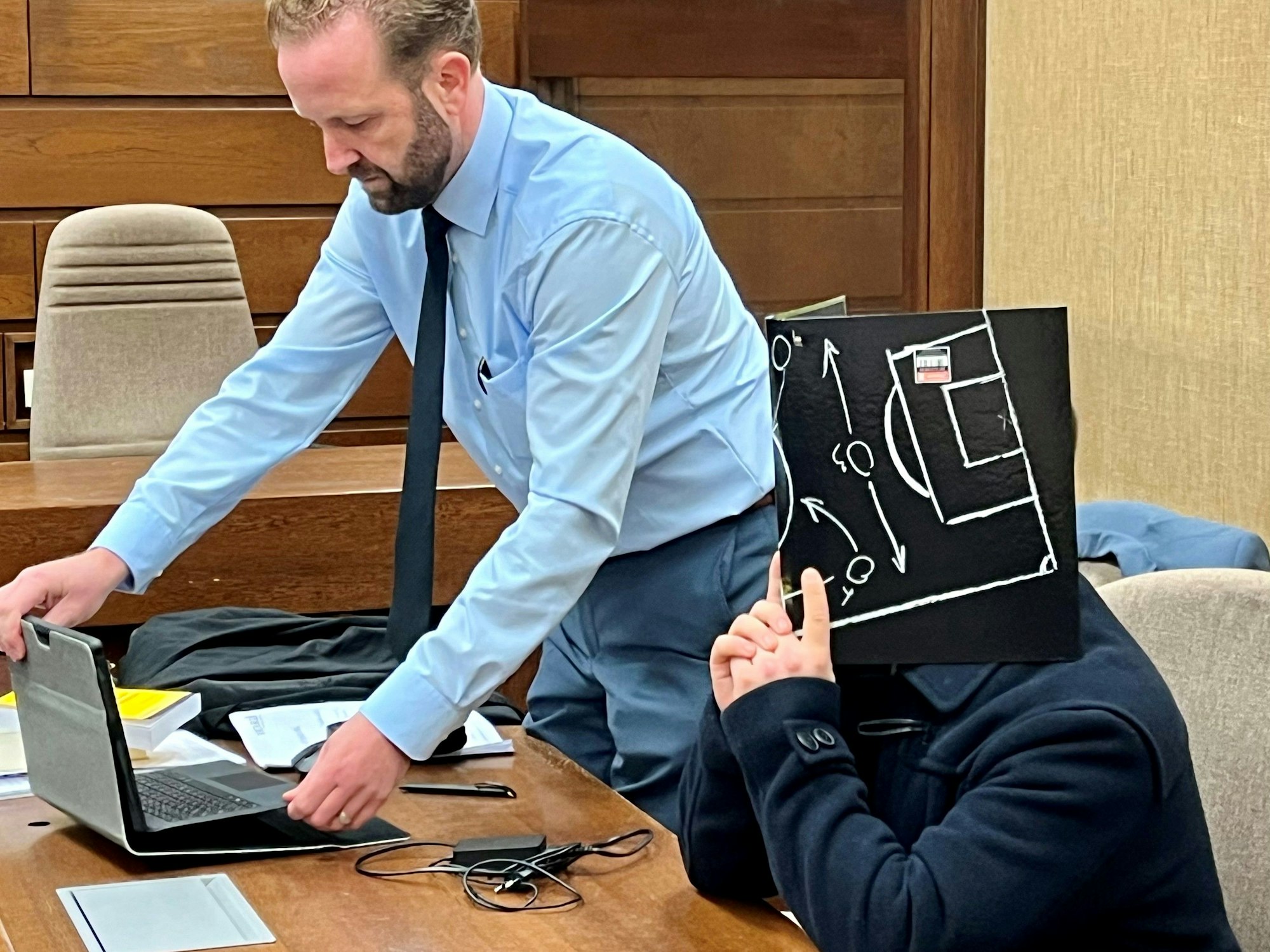 Ein Mann hält sich eine Mappe vor das Gesicht, neben ihm steht sein Anwalt.