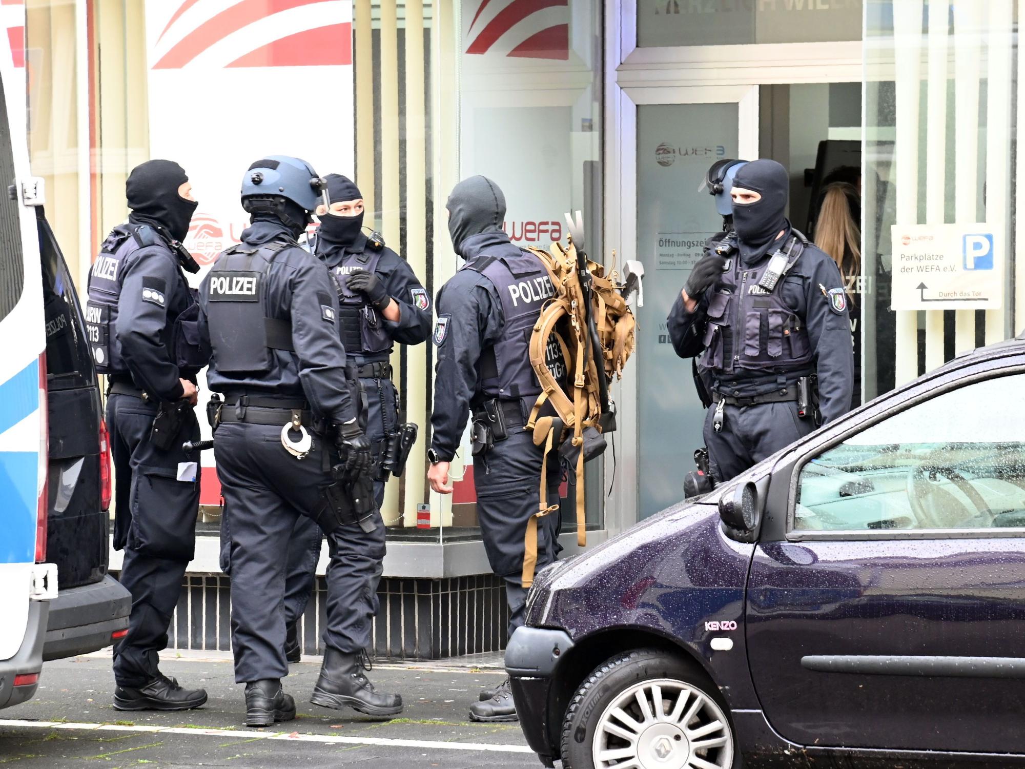 Fünf vermummte Einsatzkräfte stehen vor einer Eingangstür eines Gebäudes.