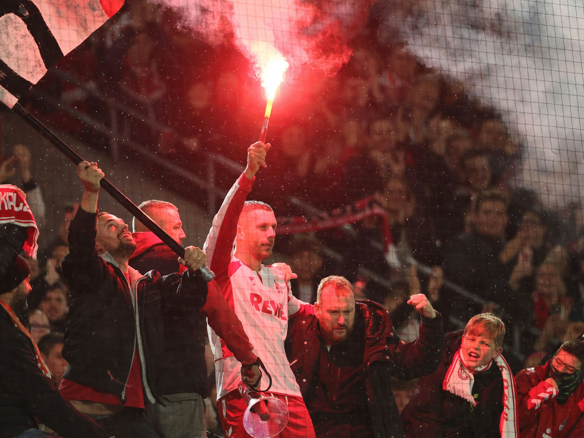 Lukas Podolski mit Pyro in der Hand bei den Fans auf dem Zaun im Stadion.