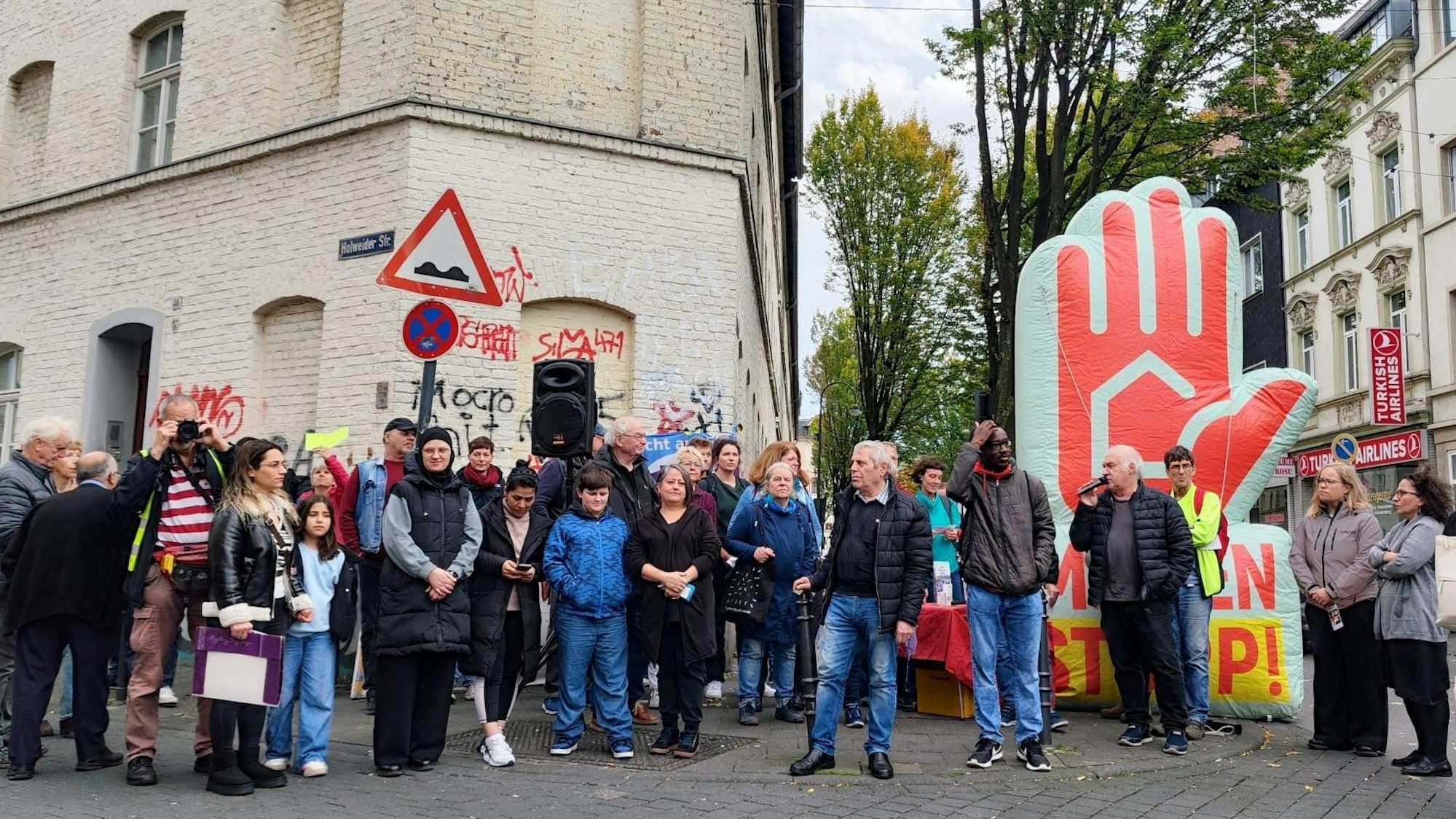 Mehrere Menschen stehen nebeneinander vor einem Haus auf der Straße. Ein Mann hält ein Mikro.