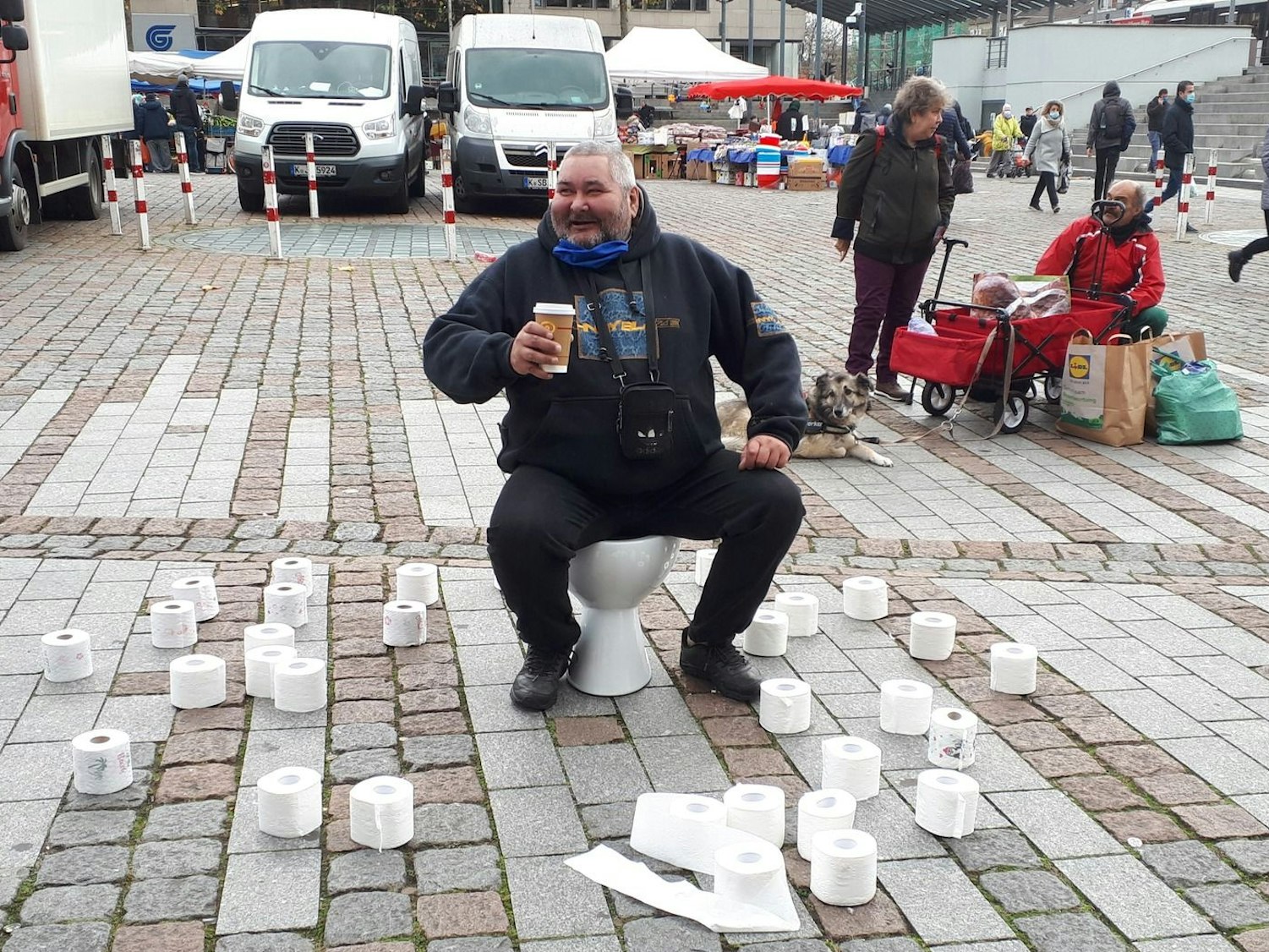 Ein Mann sitzt lachend auf einer Toilette, die auf einem Platz aufgestellt wurde und um die herum zahlreiche Klopapierrollen liegen.