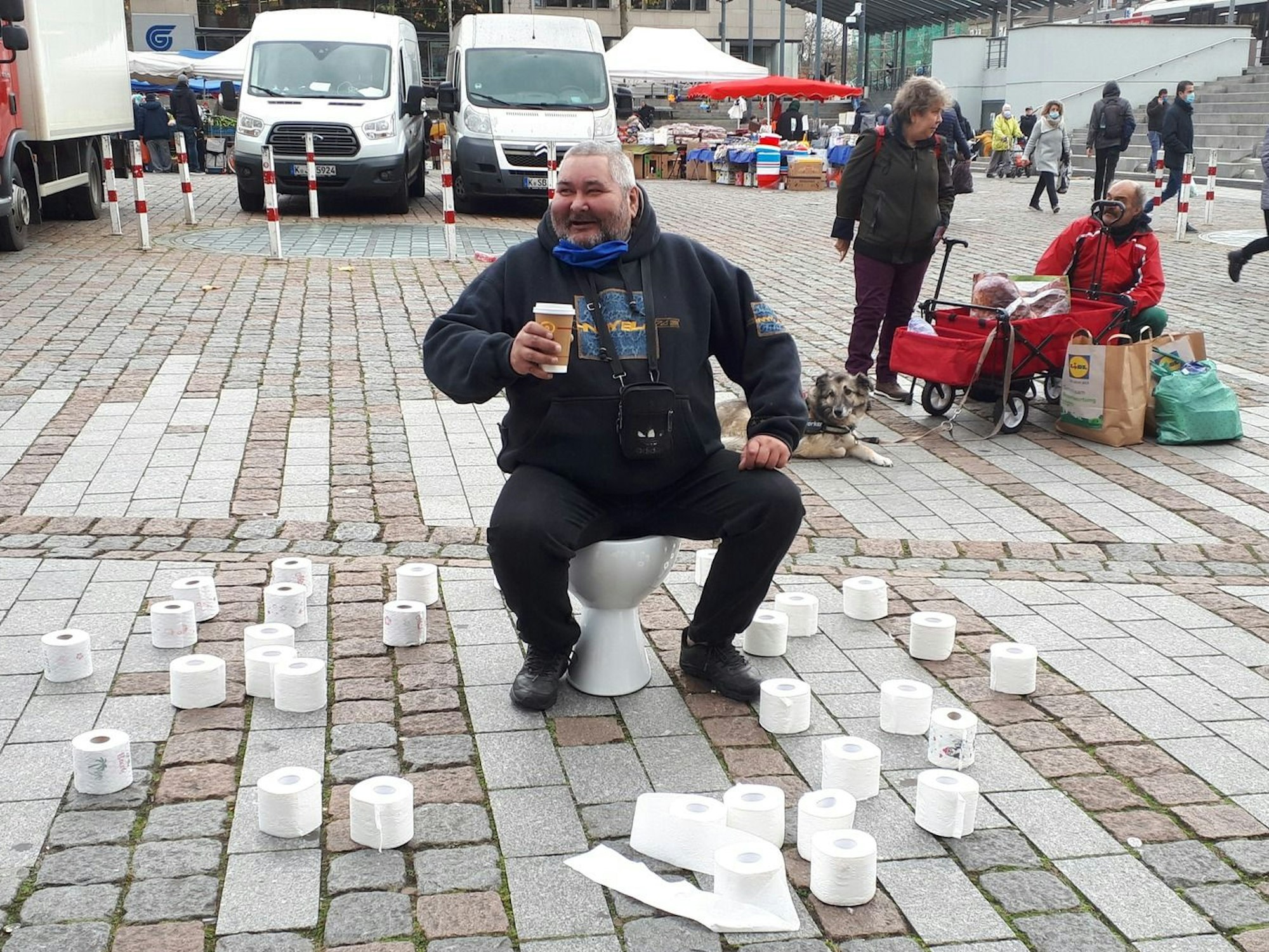 Ein Mann sitzt lachend auf einer Toilette, die auf einem Platz aufgestellt wurde und um die herum zahlreiche Klopapierrollen liegen.