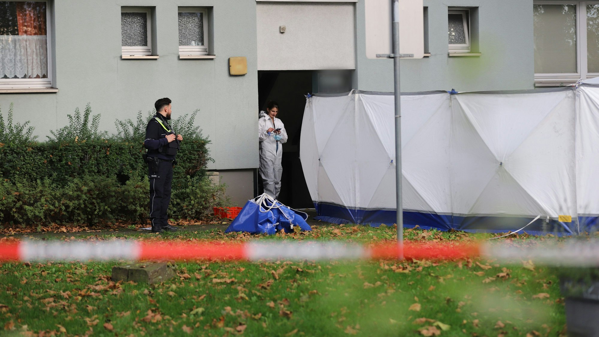 Zwei Einsatzkräfte der Polizei stehen vor einem Wohnhaus, wo auf dem Rasen ein Sichtschutz aufgebaut wurde.