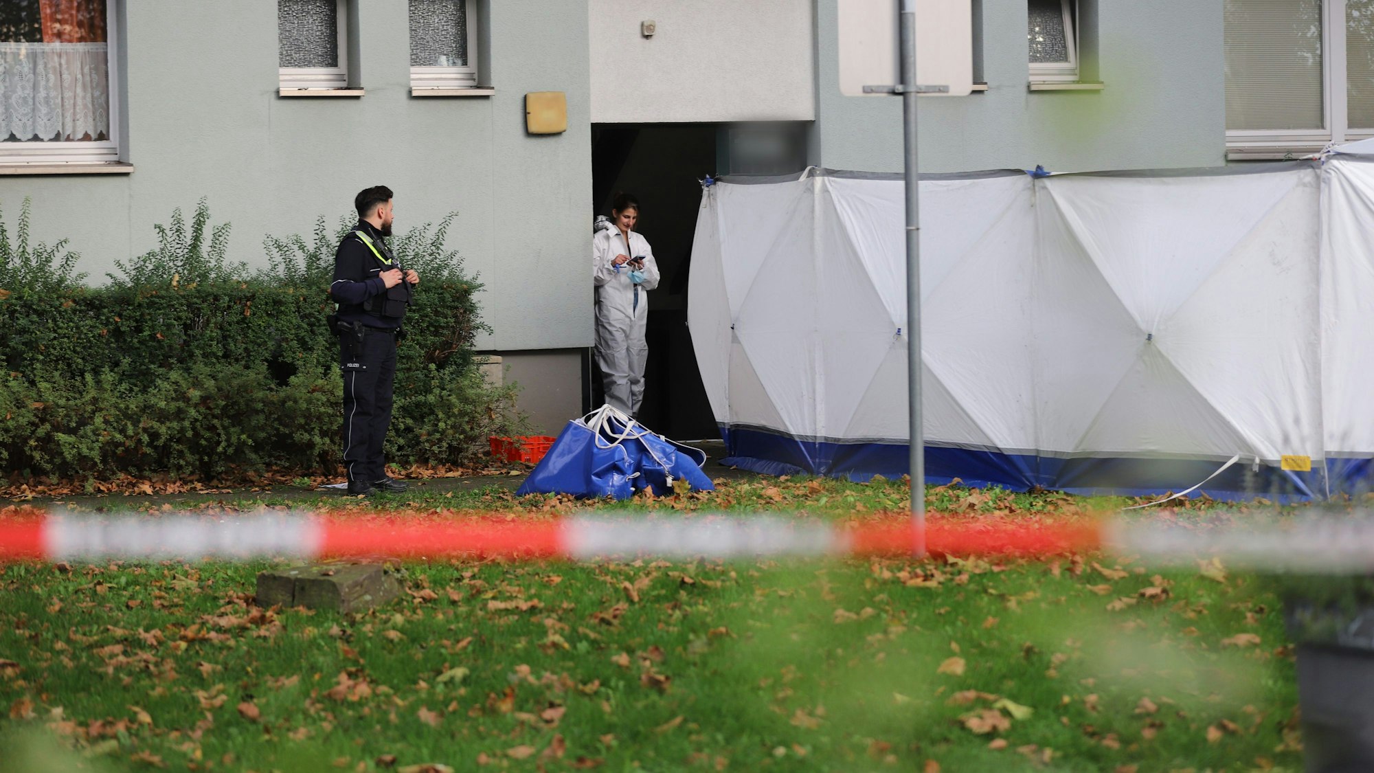Ein Polizist und eine Beamtin der Spurensicherung stehen vor einem Haus, neben ihnen ist ein Sichtschutz aufgebaut.