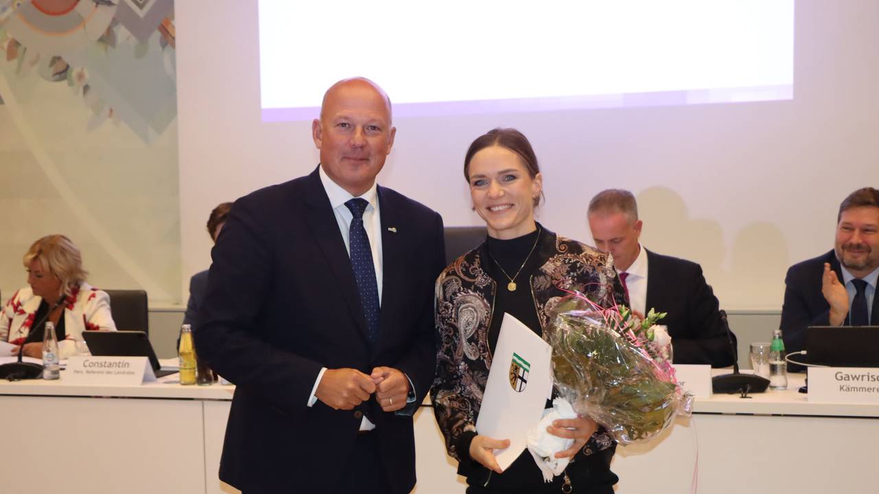 Eine Frau hält einen Blumenstrauß und eine Urkunde in der Hand, neben ihr steht ein Mann.