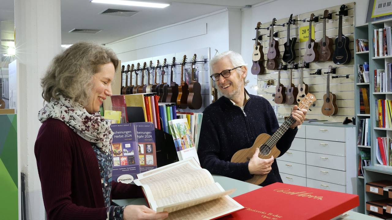 Matthias und Astrid Hinz stehen in ihrem Musikladen und grinsen sich an. Im Hintergrund sind diverse Gitarren und andere Instrumente zu sehen.