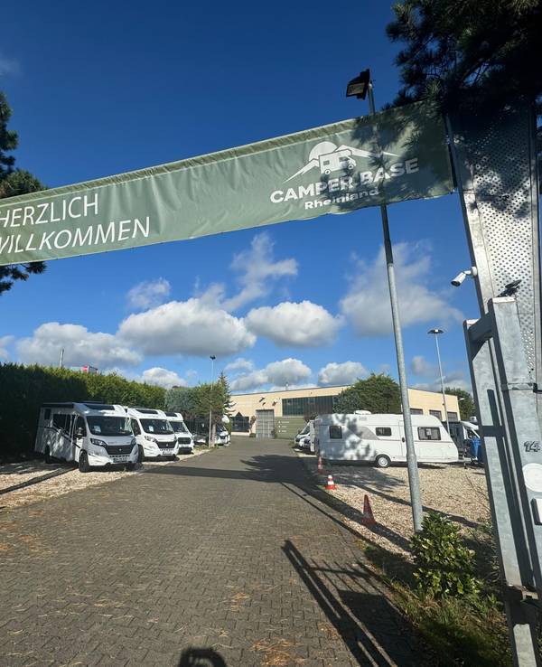 Über einem geöffneten Tor zu einem Gelände, auf dem Camping-Busse stehen, hängt ein Banner mit den Worten „Herzlich Willkommen Camper Base Rheinland“.