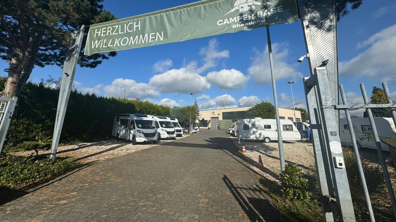 Über einem geöffneten Tor zu einem Gelände, auf dem Camping-Busse stehen, hängt ein Banner mit den Worten „Herzlich Willkommen Camper Base Rheinland“.