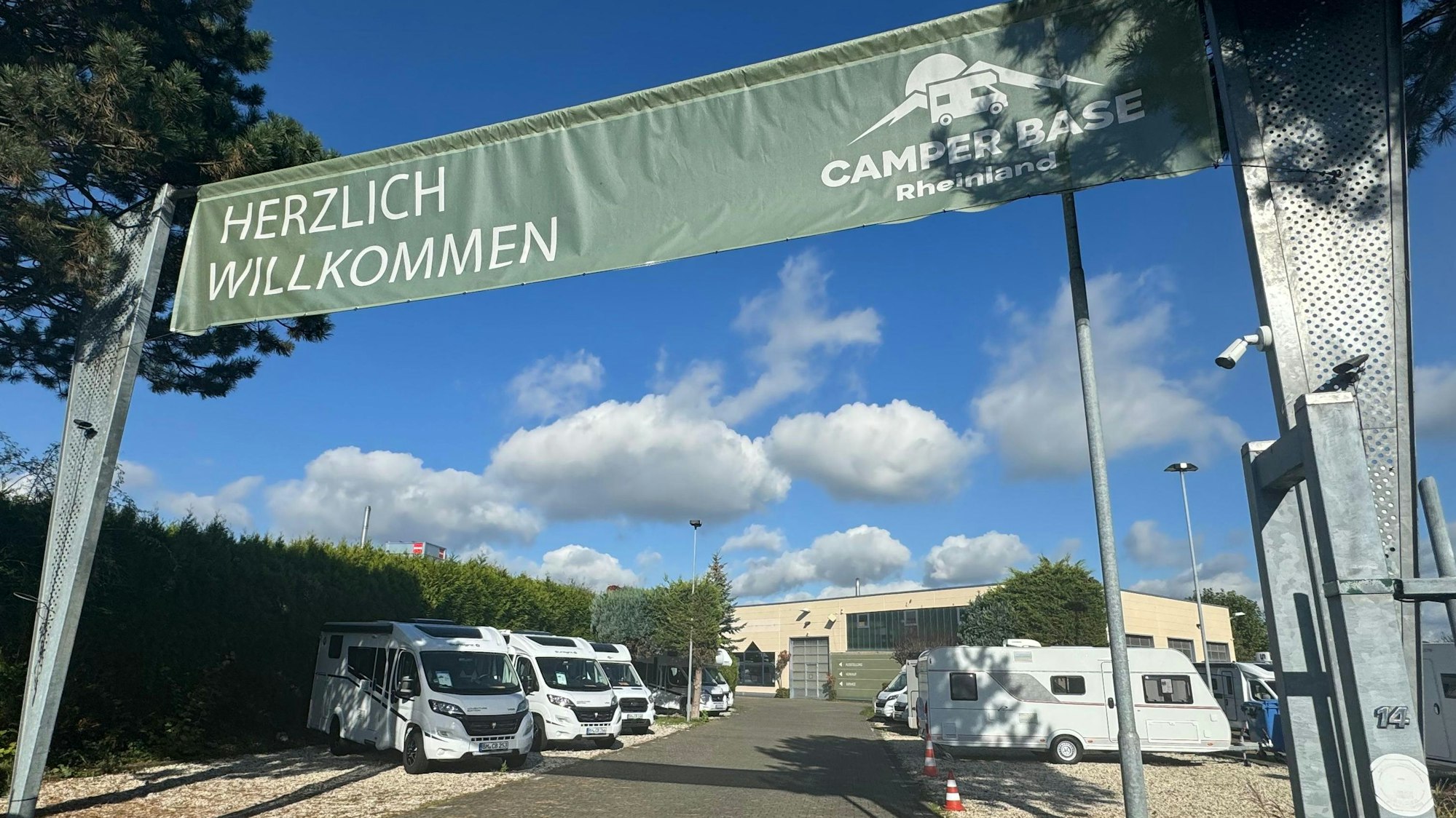 Über einem geöffneten Tor zu einem Gelände, auf dem Camping-Busse stehen, hängt ein Banner mit den Worten „Herzlich Willkommen Camper Base Rheinland“.