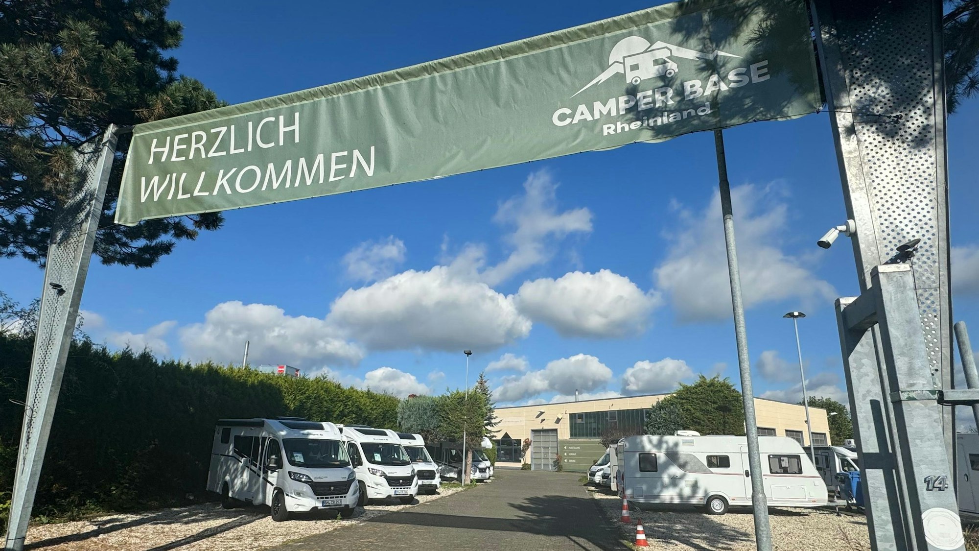 Über einem geöffneten Tor zu einem Gelände, auf dem Camping-Busse stehen, hängt ein Banner mit den Worten „Herzlich Willkommen Camper Base Rheinland“.