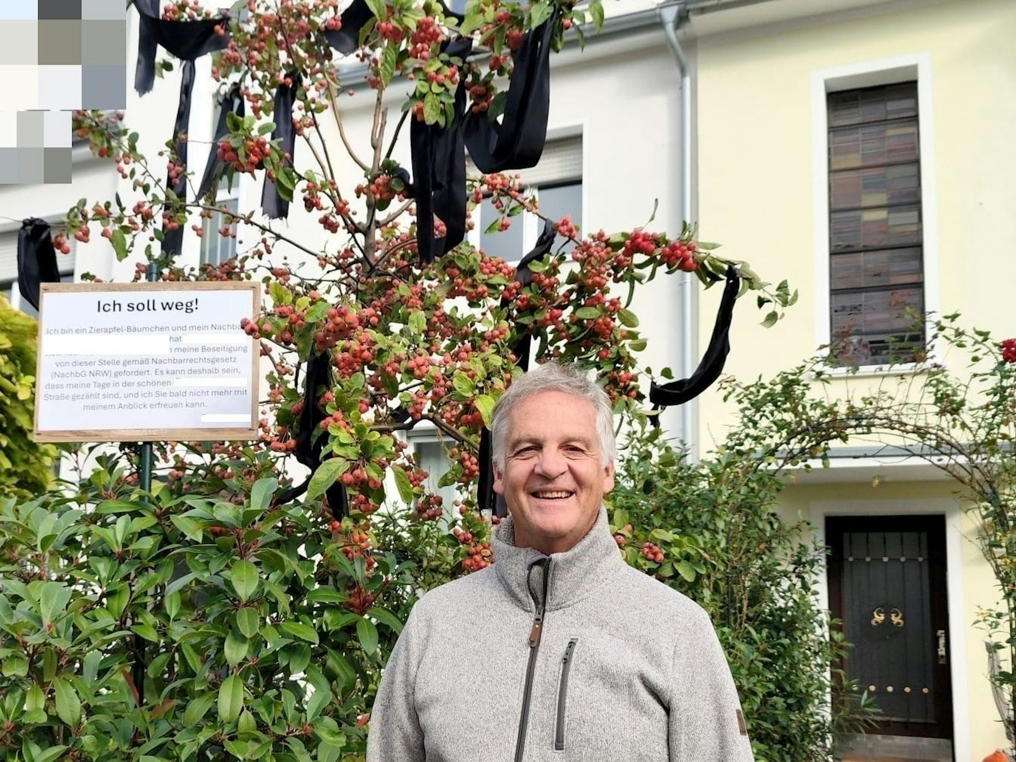 Ein Mann steht vor einem Zierapfelbaum, in dem Trauerflor und ein Schild hängen.