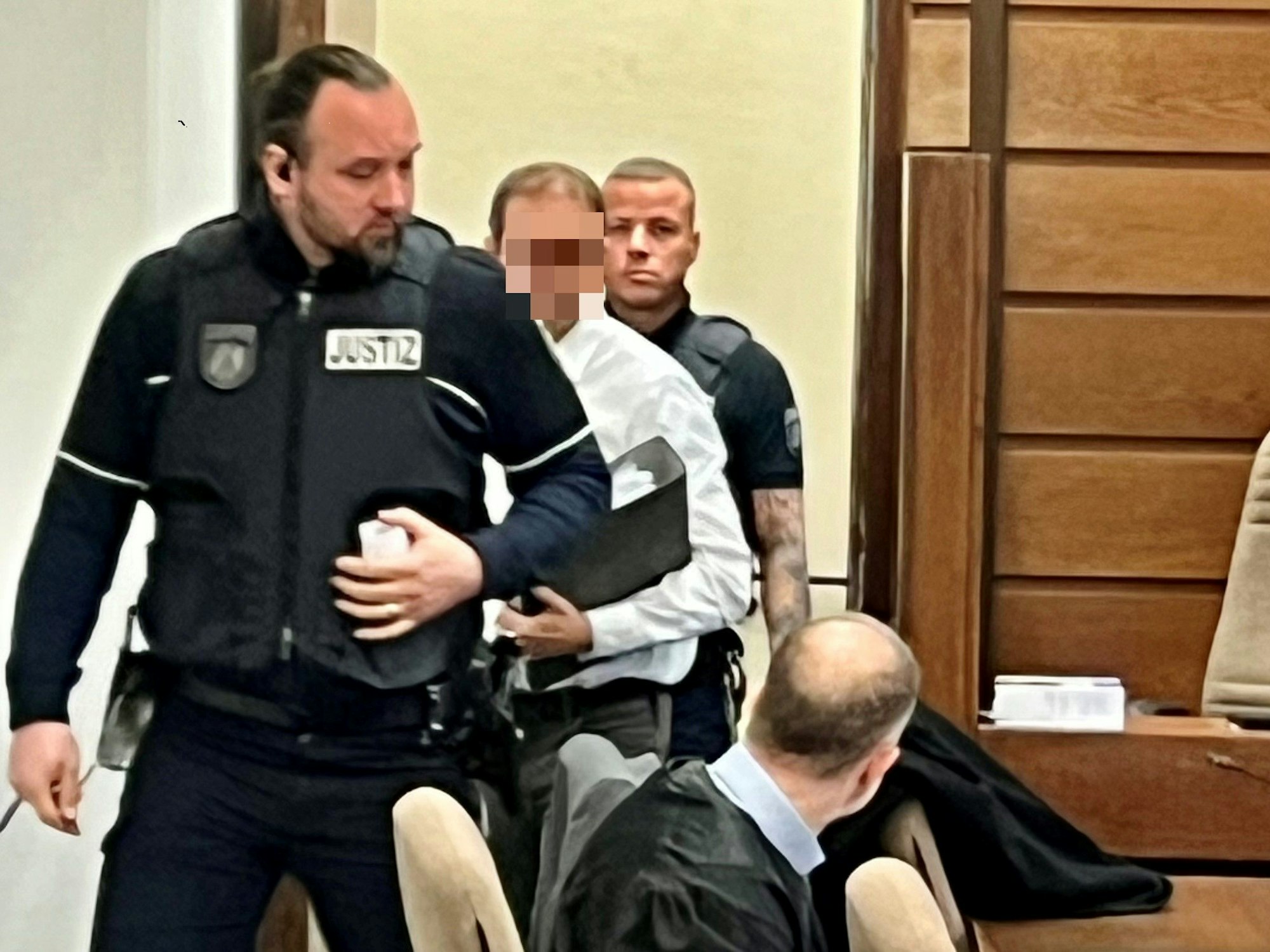 Ein Mann betritt mit zwei Justizwachtmeistern einen Gerichtssaal, unter seinem Arm hat er eine Akte.