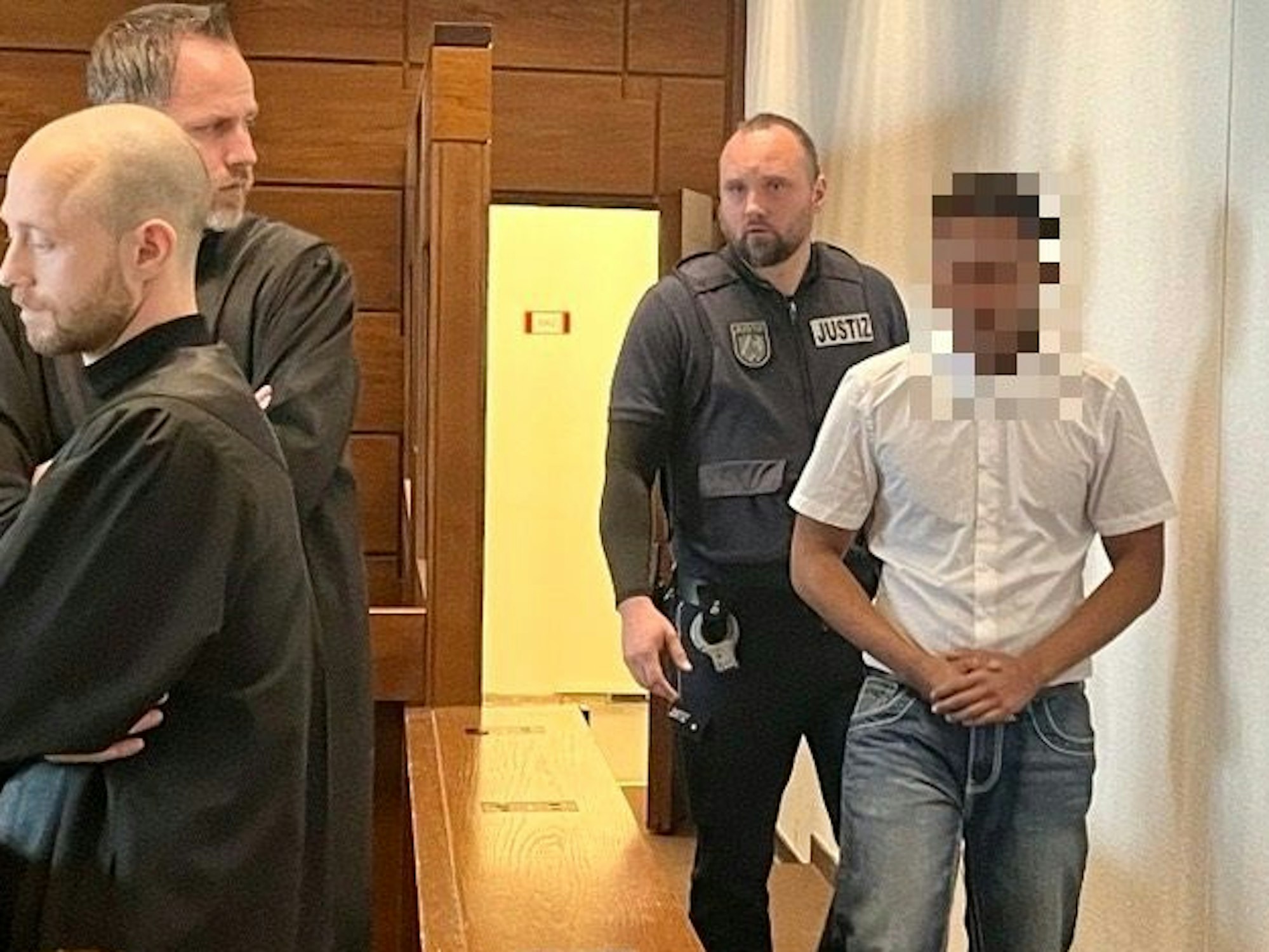 Ein dunkelhäutiger junger Mann wird von einem Justizwachtmeister in den Gerichtssaal gebracht. Ein Anwalt dreht sich zu ihm um.