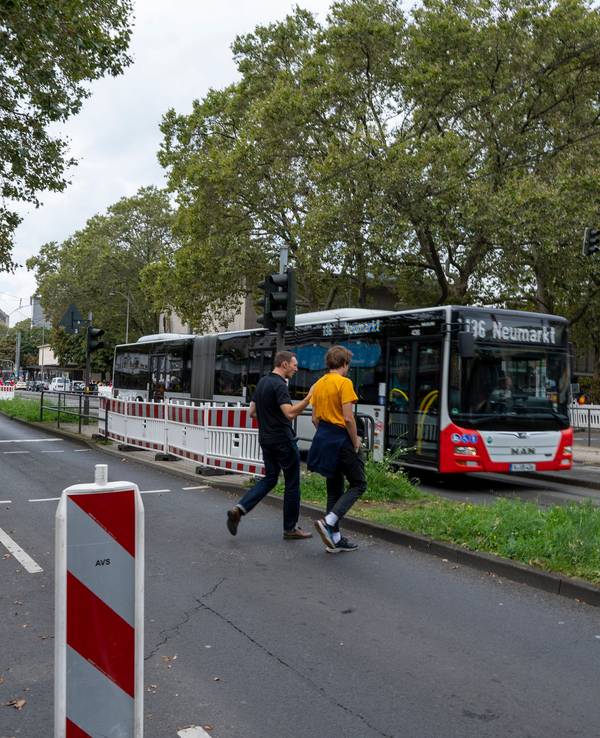 Zwei Männer gehen über eine Ampel, im Hintergrund sind Autos und ein Bus auf der Straße zu sehen.