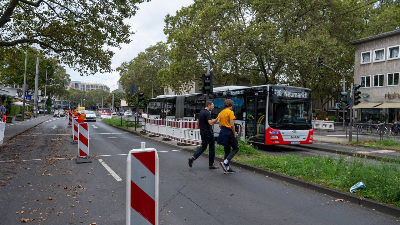 Zwei Männer gehen über eine Ampel, im Hintergrund sind Autos und ein Bus auf der Straße zu sehen.
