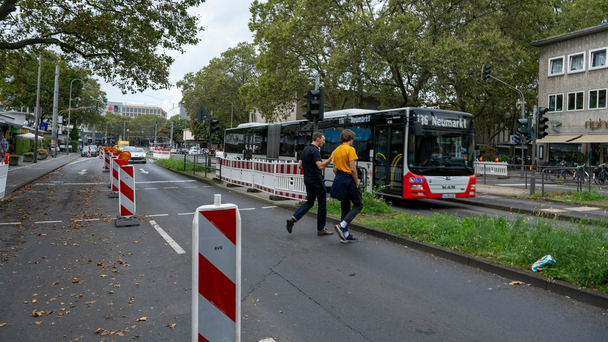 Zwei Männer gehen über eine Ampel, im Hintergrund sind Autos und ein Bus auf der Straße zu sehen.