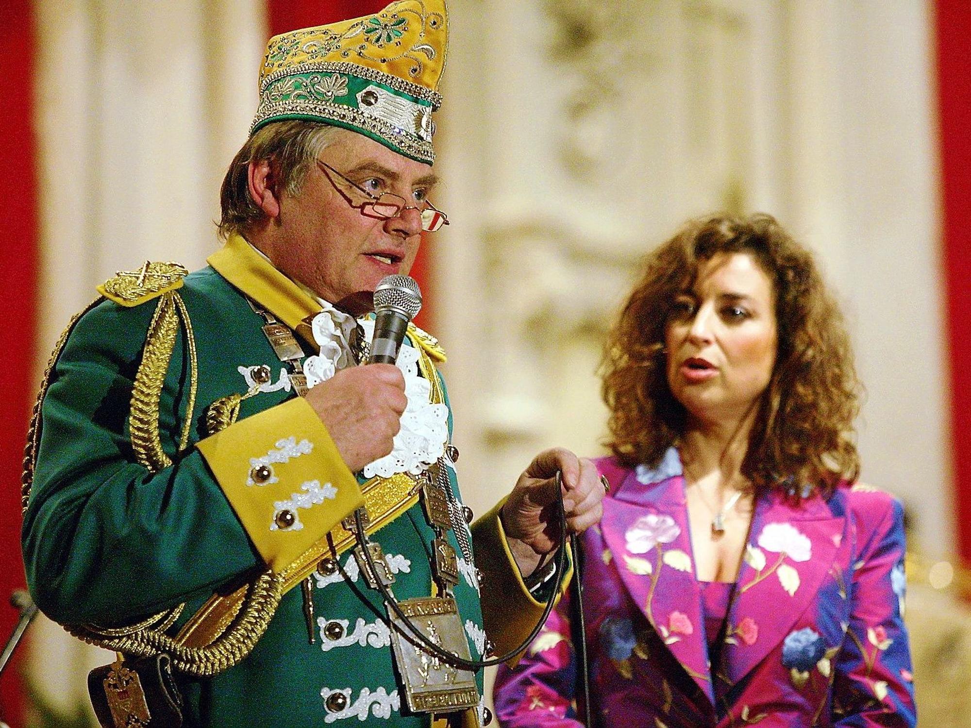 Reiner Benner spricht ins Mikrofon zum Saal, im Hintergrund die konsternierte Isabel Varell.