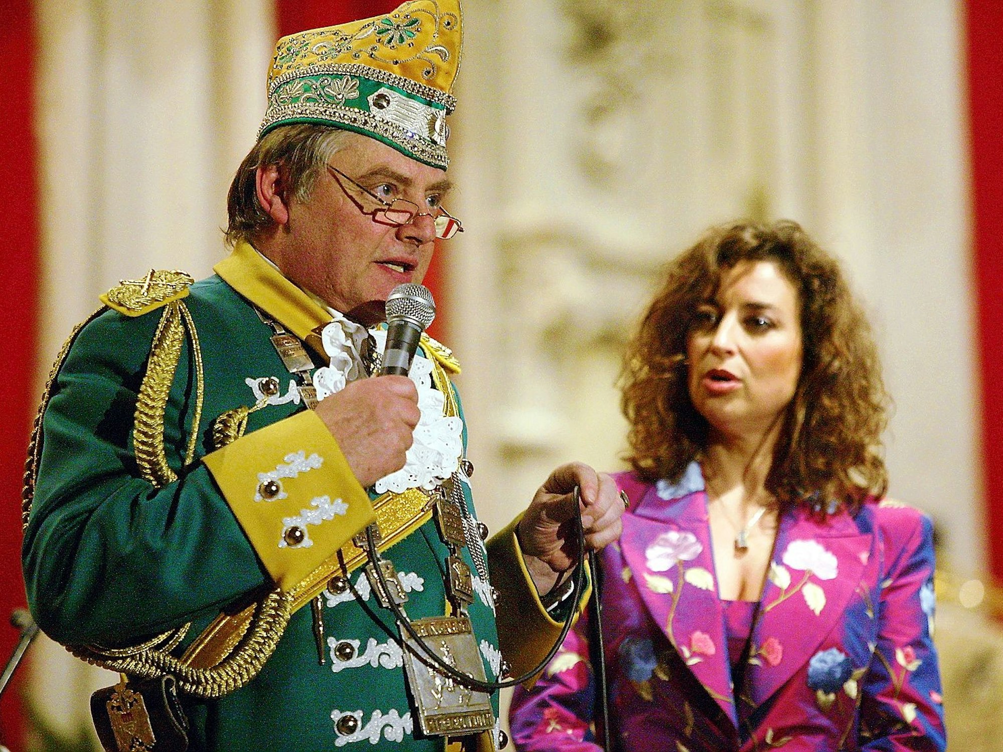 Reiner Benner spricht ins Mikrofon zum Saal, im Hintergrund die konsternierte Isabel Varell.