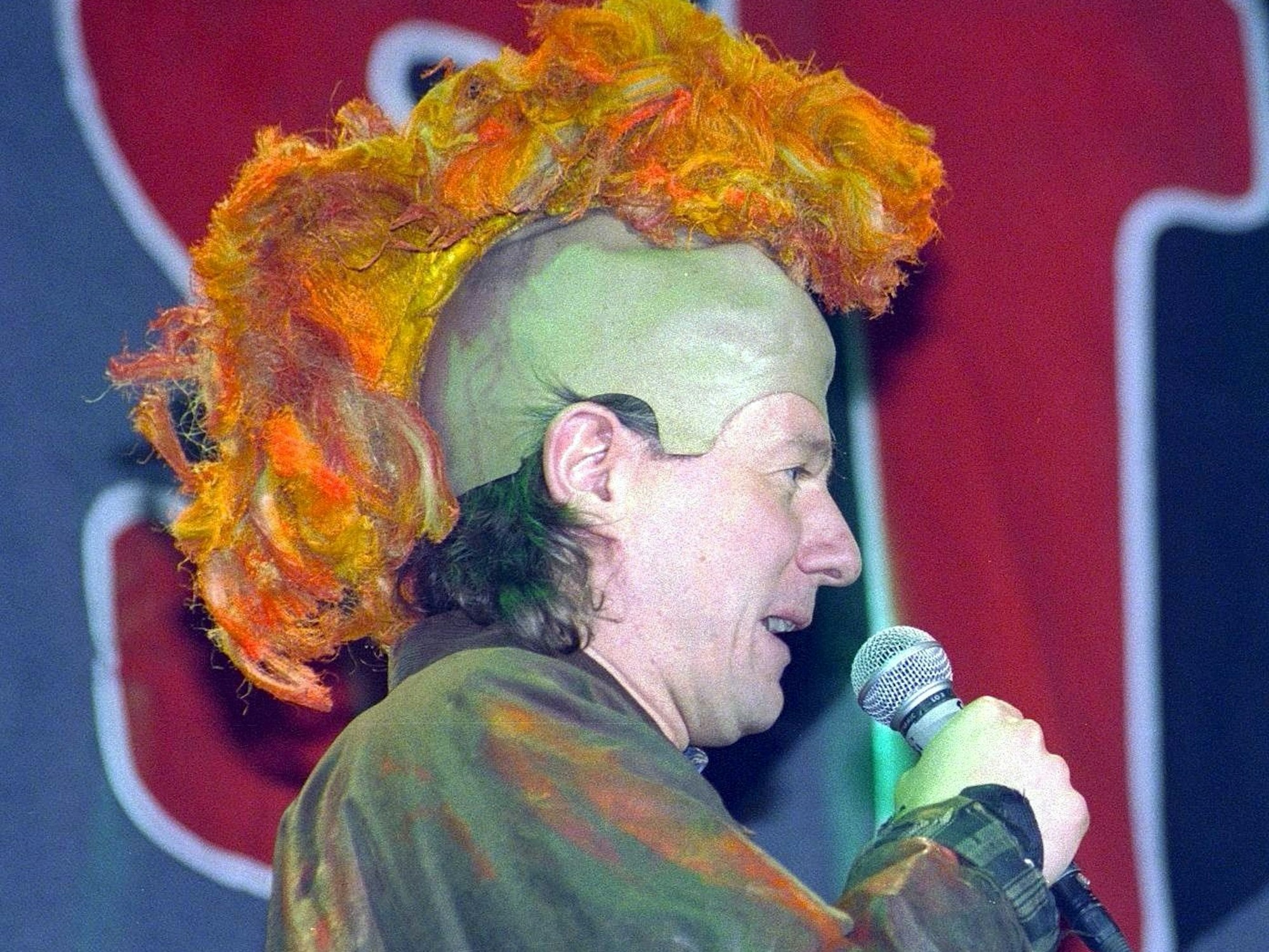 Jürgen Becker hat eine Irokesen-Perücke auf und spricht ins Mikrofon.