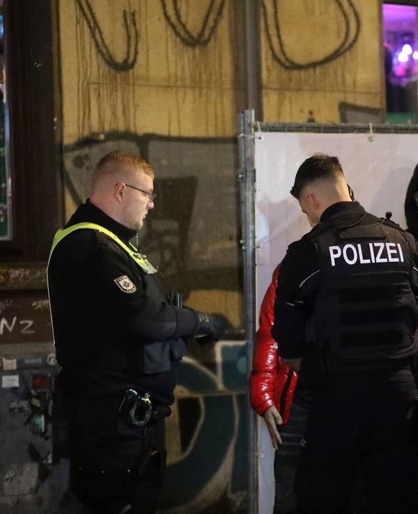 Ein Polizist kontrolliert einen Jugendlichen, der neben weiteren Jugendlichen an einer Absperrung steht. Neben dem Polizisten stehen zwei Kräfte des Ordnungsamtes.