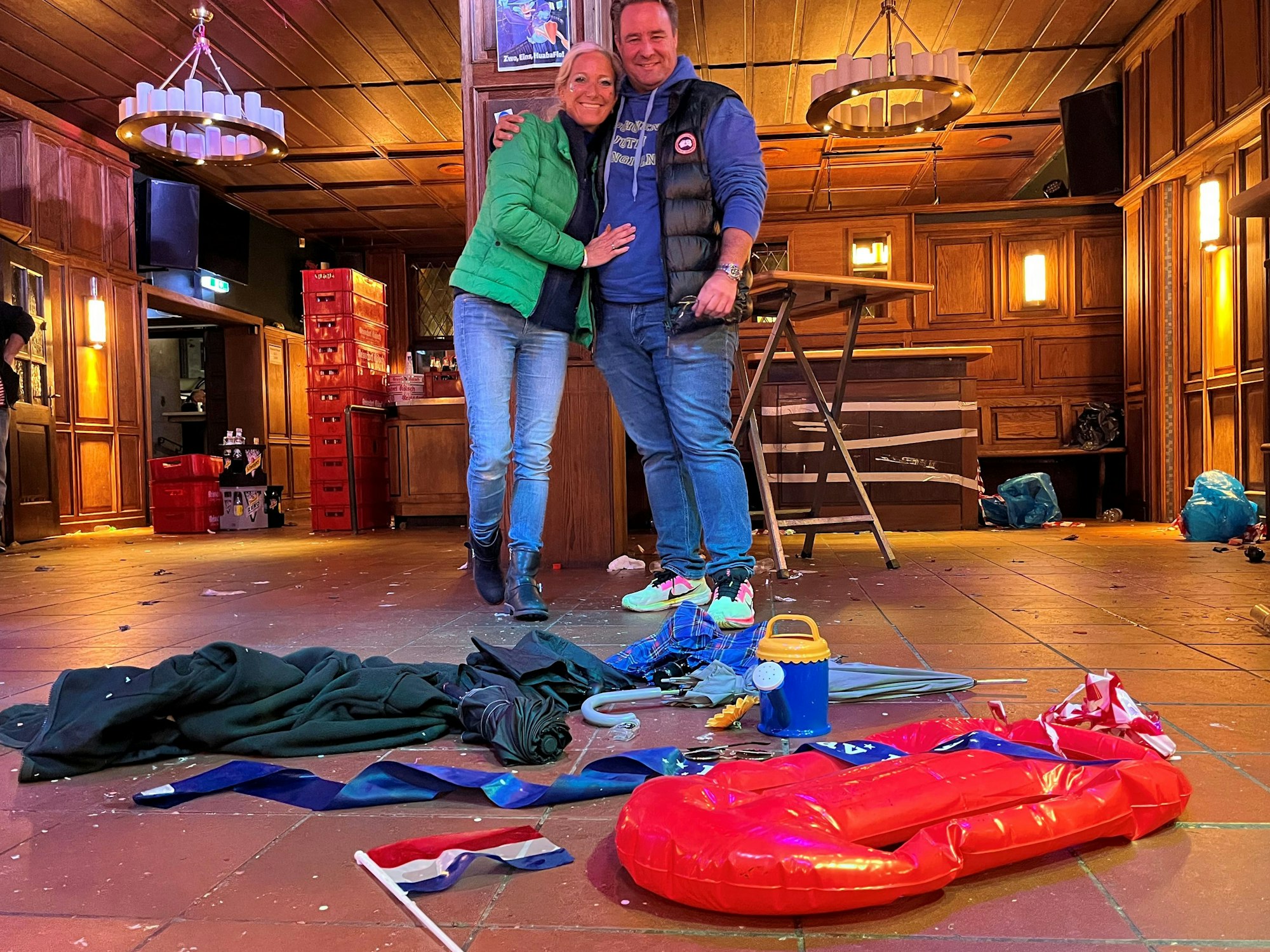 Ein Paar steht in einem Kneipenraum, auf dem Boden liegen unter anderem eine Plastikgießkanne, ein Regenschirm und eine Fahne.