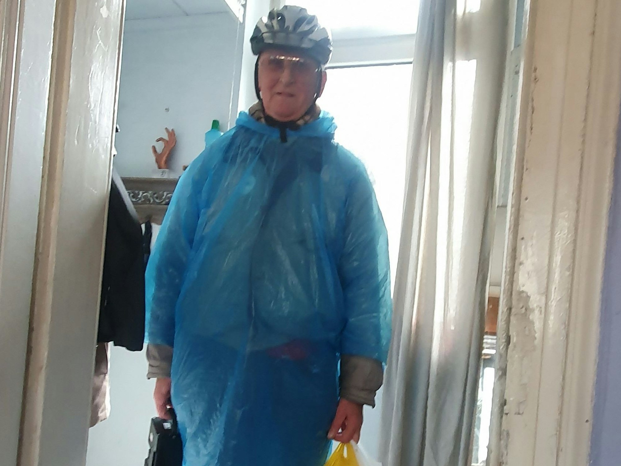 Ein älterer Mann steht in einer geöffneten Tür, er trägt einen Fahrradhelm und einen Regenschutz aus Plastik.