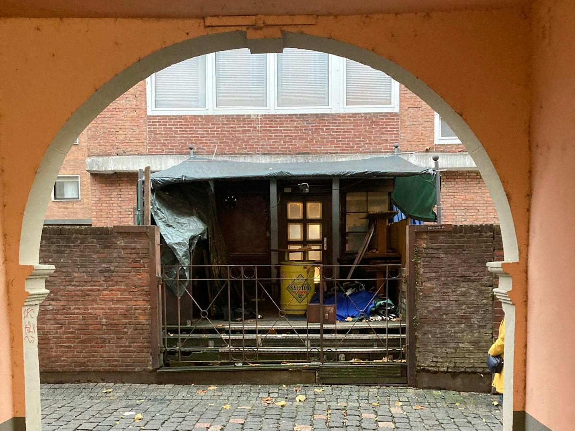 Durch einen Torbogen blickt man auf den Hinterausgang eines Gebäudes, vor dem Möbel gestapelt sind und Gerümpel auf dem Boden liegt.