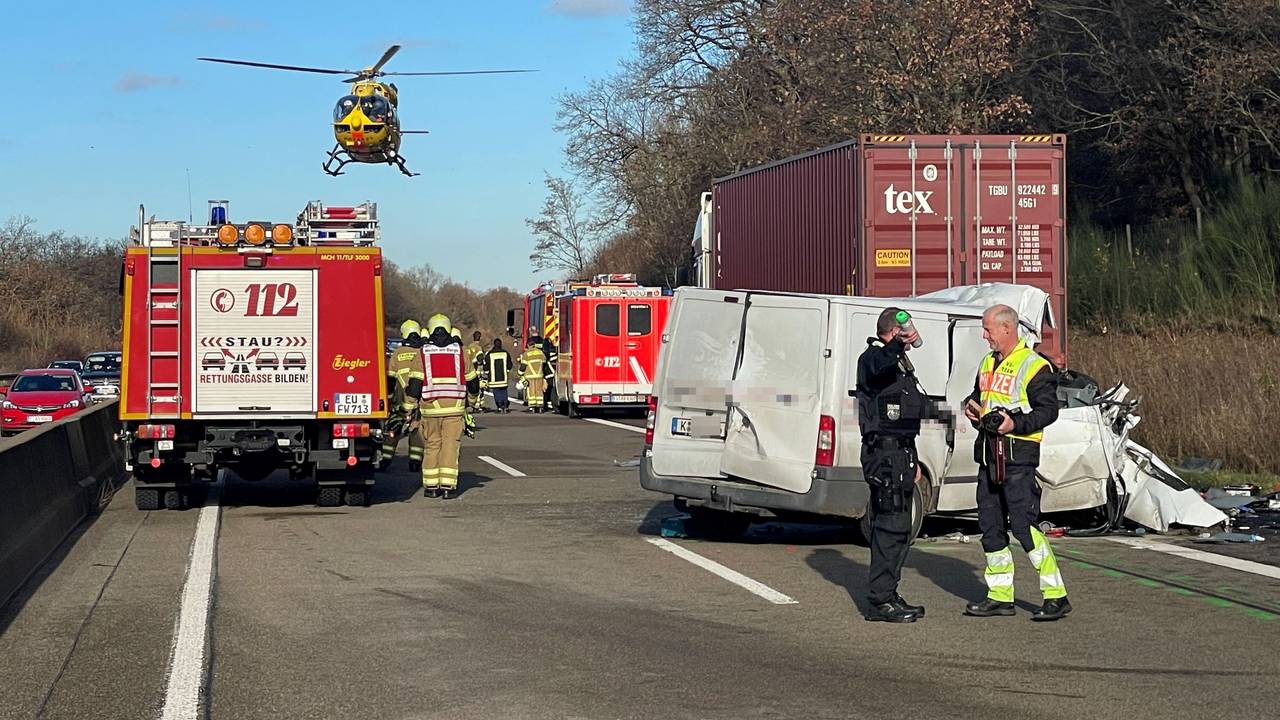 Nach einem Unfall steht ein stark beschädigter Transporter auf einer Autobahn quer zur Fahrbahn, auf dem Standstreifen ein Lkw. Einsatzfahrzeuge und -kräfte von Feuerwehr und Polizei befinden sich auf der Autobahn, ein Hubschrauber ist in der Luft.