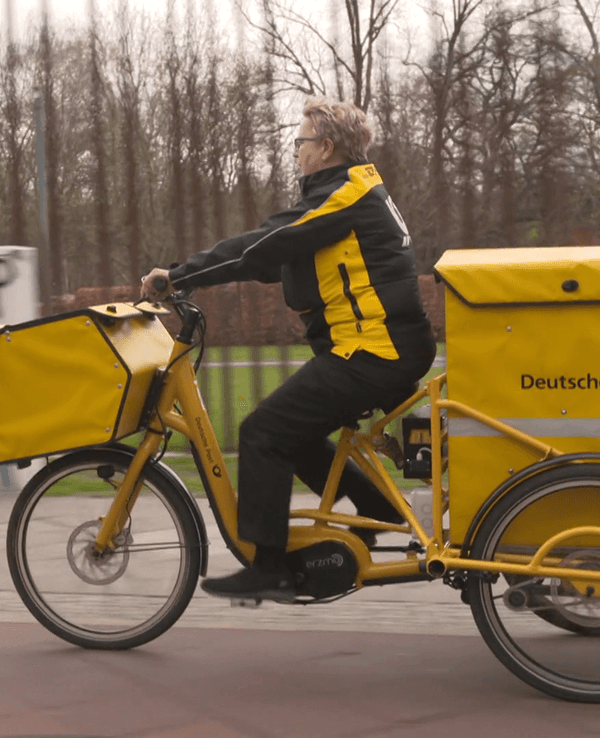 Eine Postbotin transportiert Lieferungen auf einem Fahrrad. Screenshot aus Presse-Video aus der DHL-Mediathek (erstellt von Laura Wenzel)